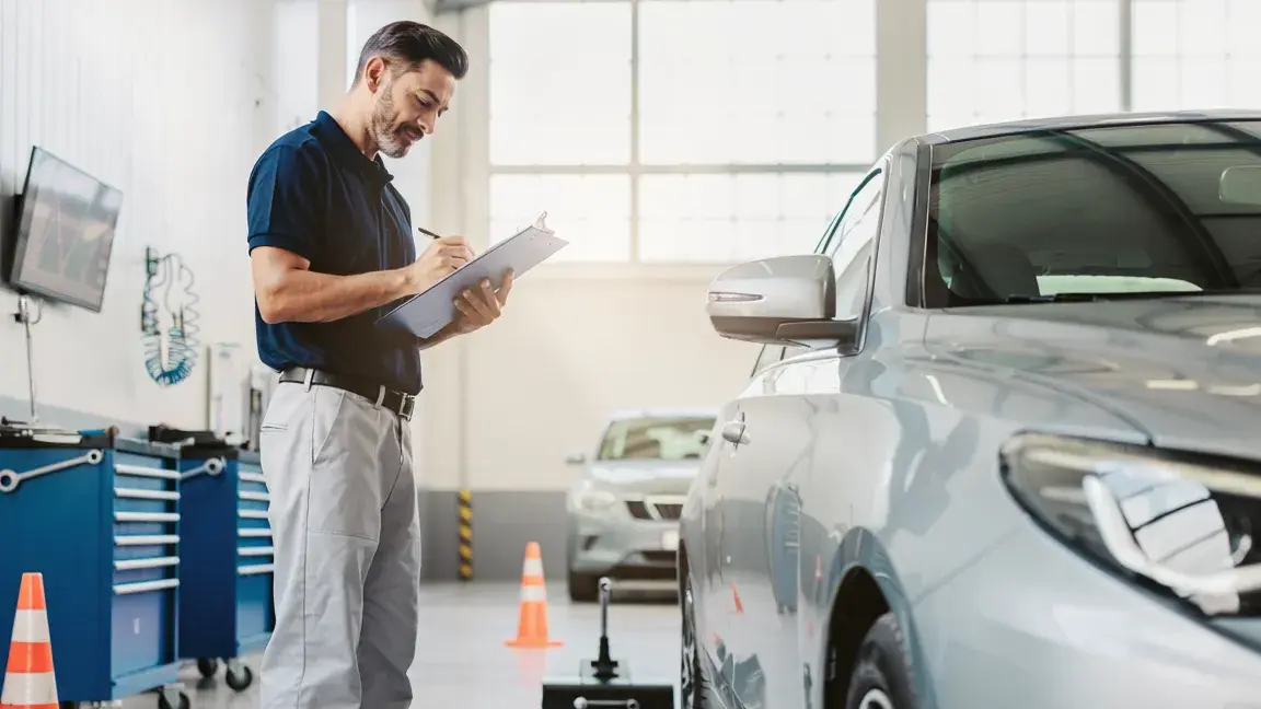 Auton kuntotutkimusta tekevä ammattilainen kirjoittaa havaintoja lehtiölle autokorjaamossa hopeisen auton vieressä.